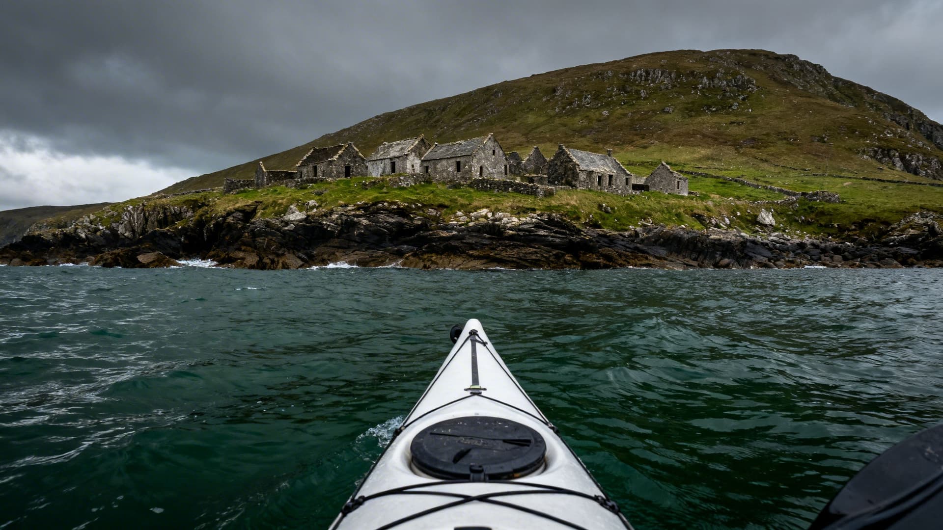 A sea kayak on the Blasket Sound, the deserted stone ruins of Great Blasket Island visible on the hillside across the water, dramatic overcast Atlantic sky, the water dark green in the foreground