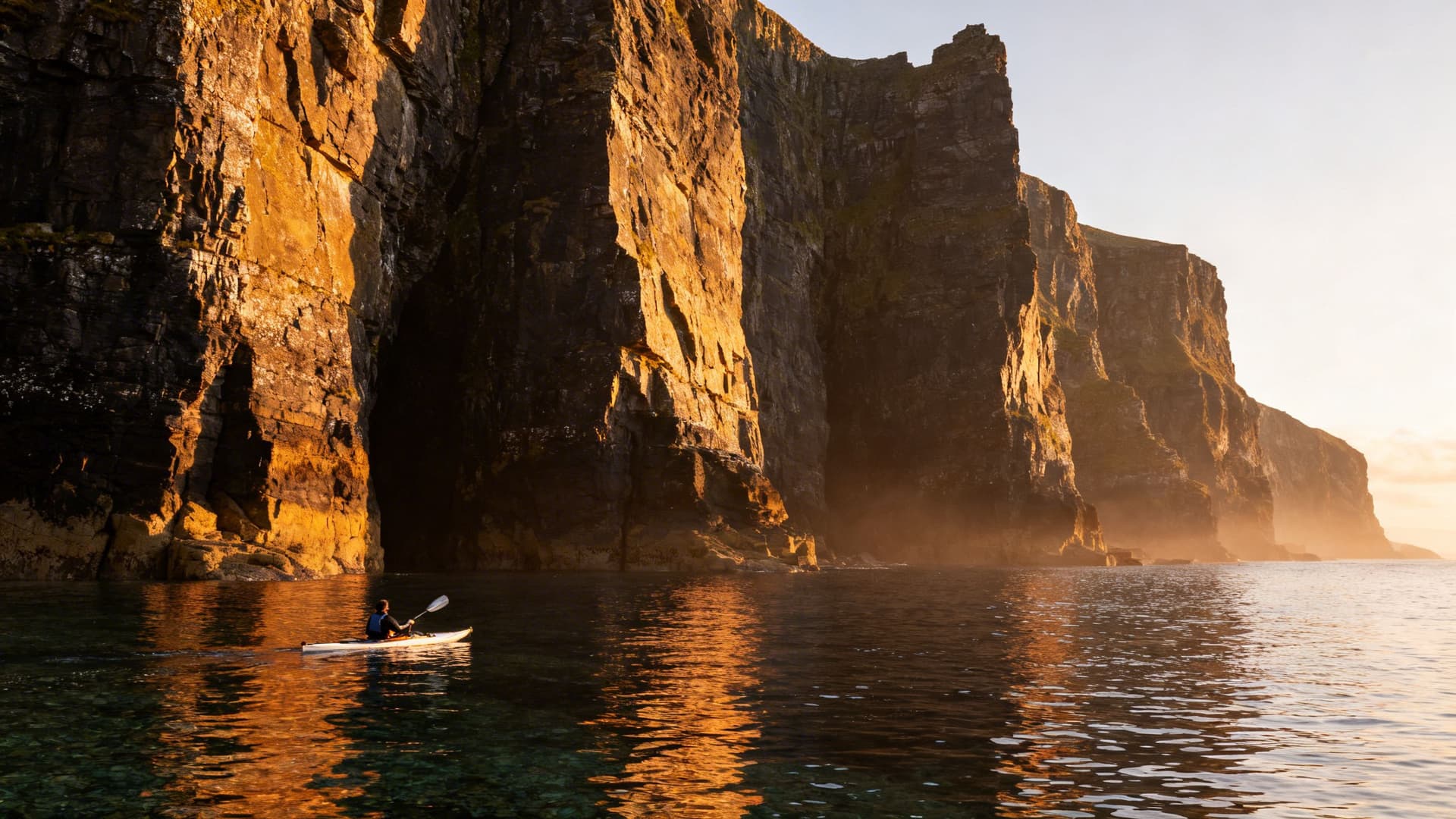 A sea kayaker paddling at the base of tall dark sea cliffs on the south shore of the Dingle Peninsula, the rock face rising dramatically above, clear Atlantic water below, late afternoon light on the stone