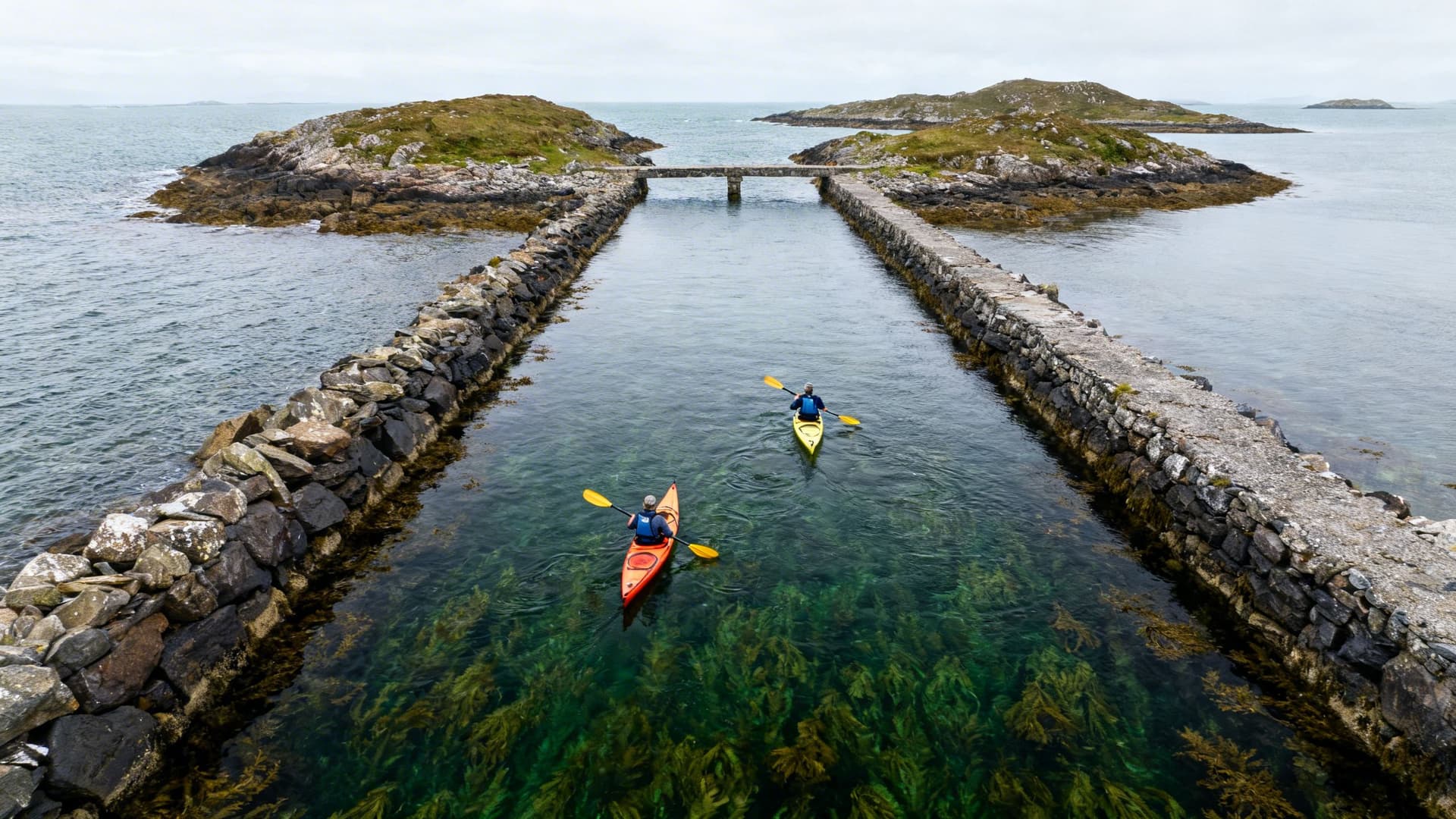 Two kayakers paddling through a narrow channel between uninhabited Connemara islands, dark green water and pale sky