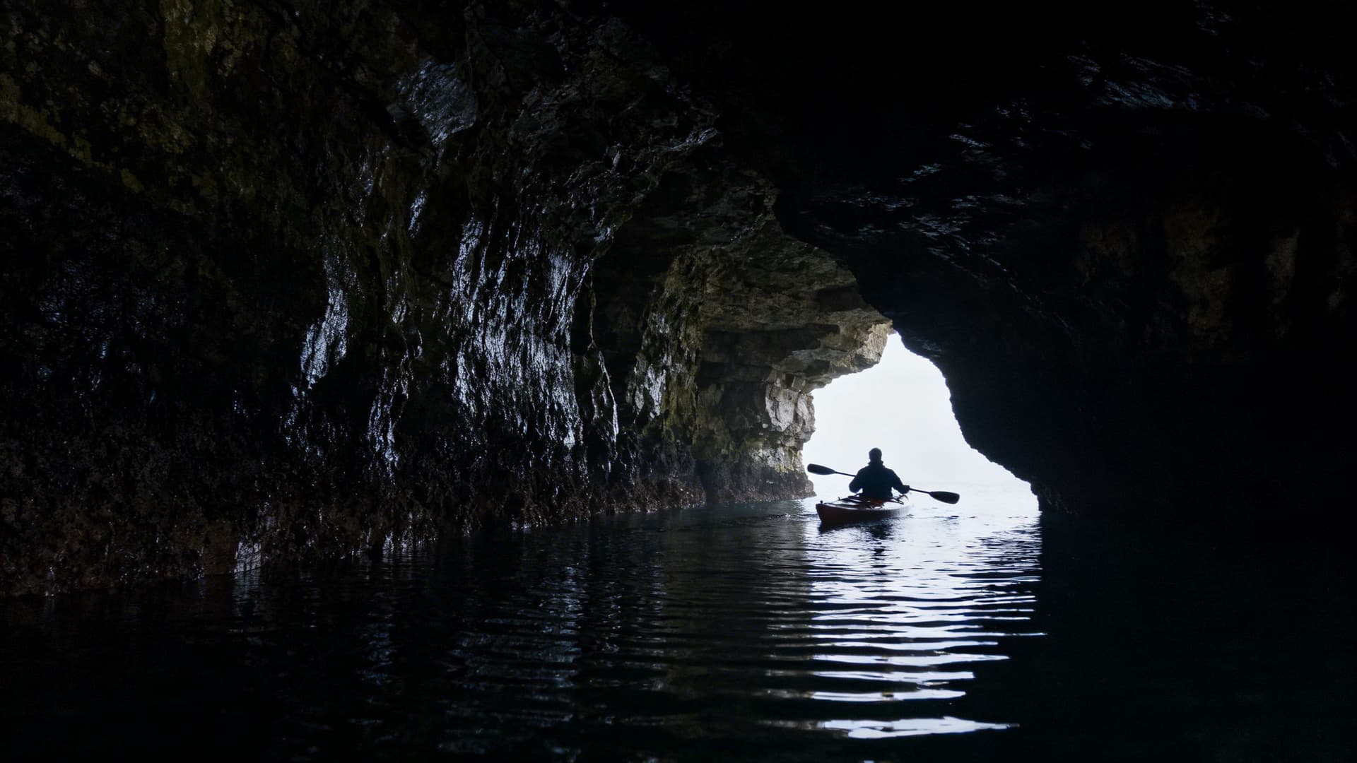 Sea kayaker inside one of the Maghera cave chambers near Ardara in County Donegal, the cave walls dark with moisture and Atlantic swell gently lifting the hull