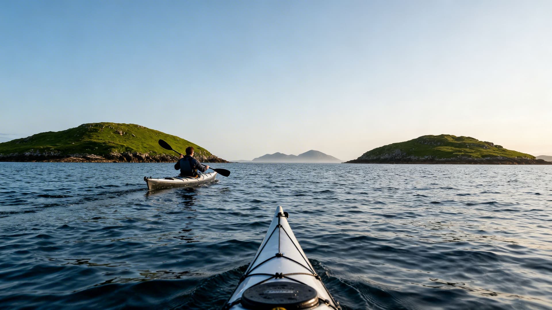 Two sea kayakers paddling toward a small uninhabited island off the Connemara coast, the Twelve Bens mountains visible in the background across calm Atlantic water