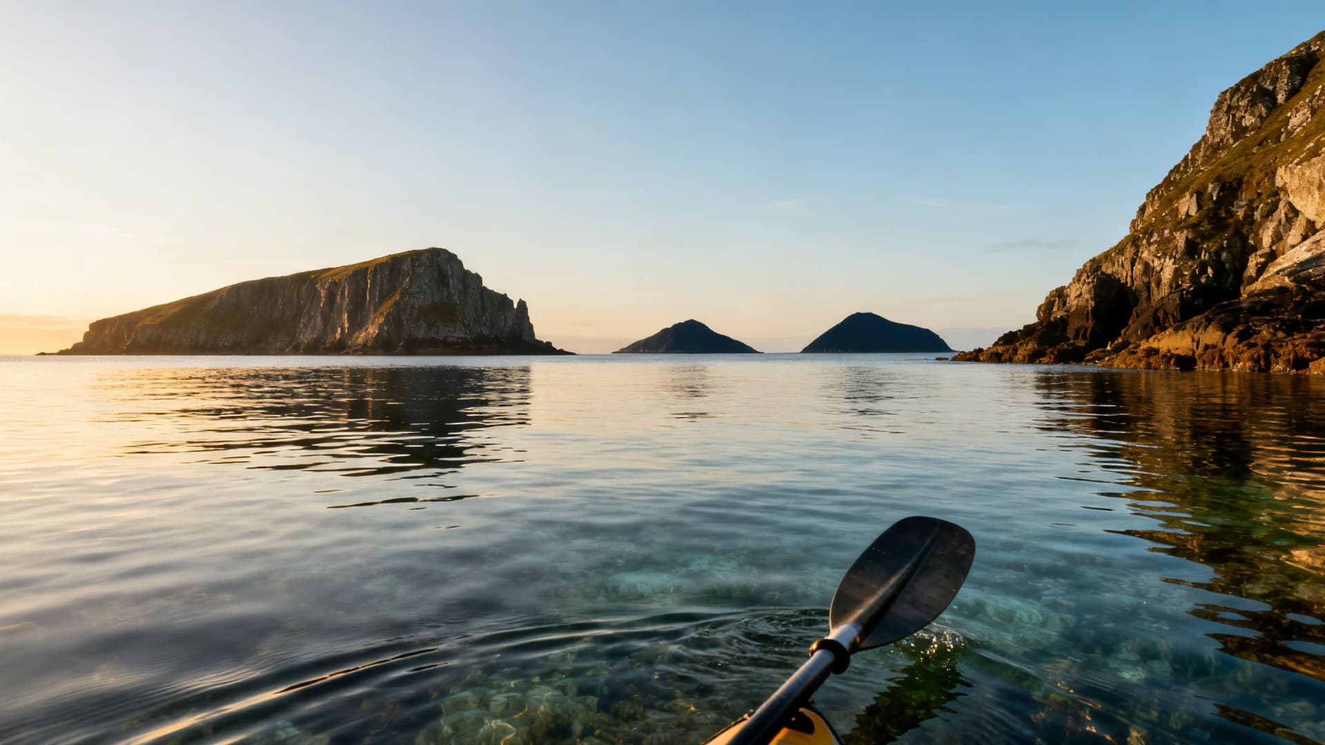 Looking west across open Atlantic water toward the silhouettes of offshore Irish islands at dawn, a sea kayaker visible at distance navigating the crossing